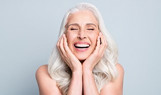 Woman with long white hair smiling with eyes closed holding her face